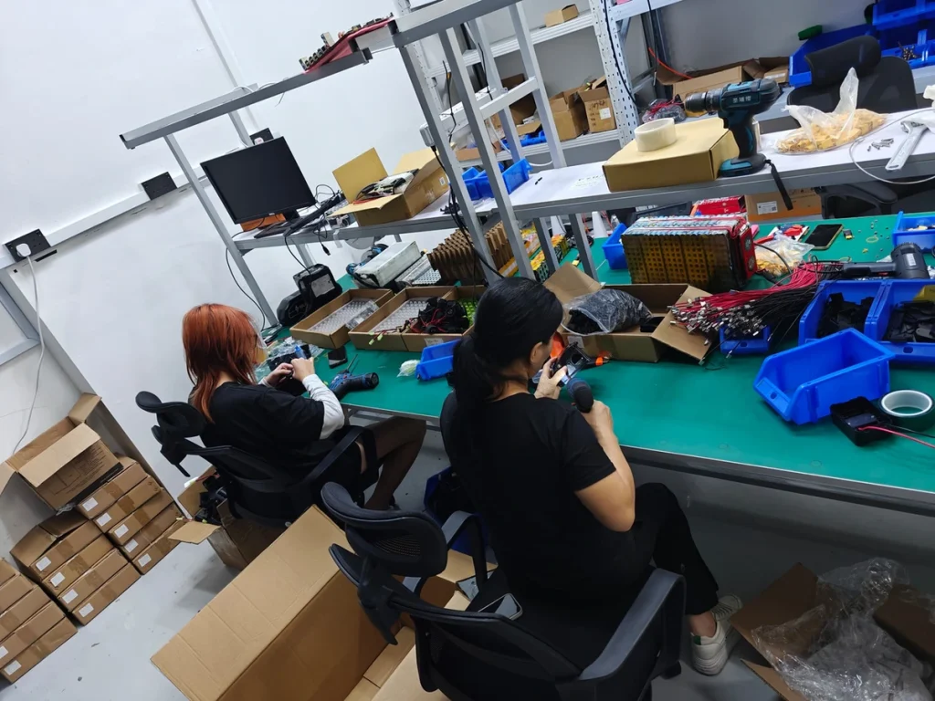 Two team members working side by side at a workbench with parts bins and boxes.
