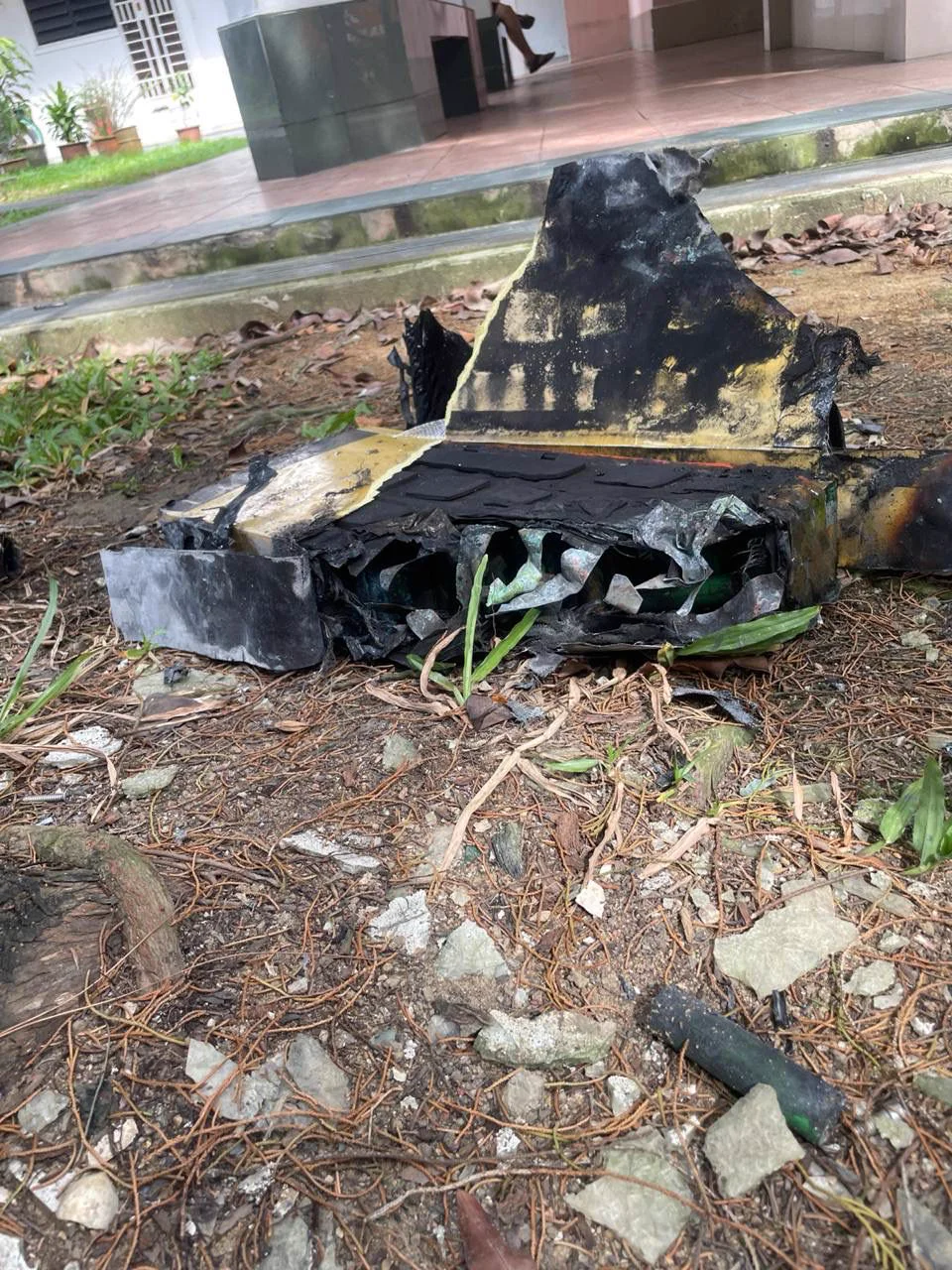 The burnt shell of a battery pack lying on soil and leaves on a grass patch