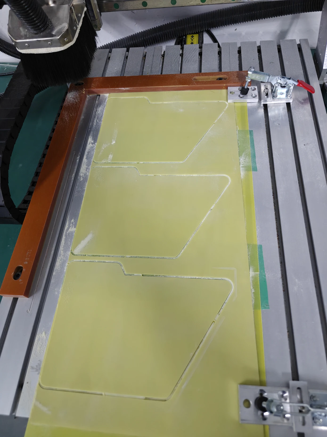A CNC router bed with shaped panels partly cut from a yellow sheet held by toggle clamps.