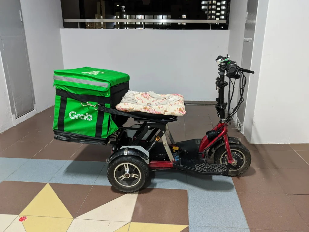 A three-wheeled electric mobility device with a green delivery bag.