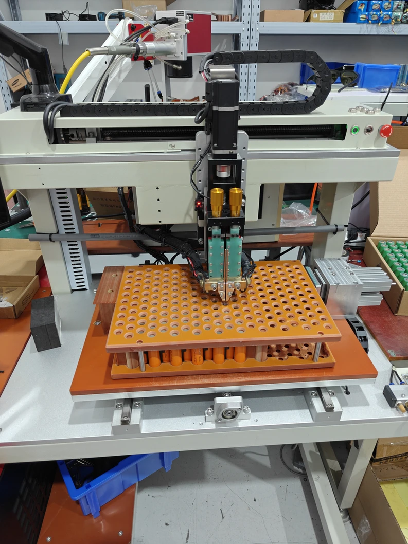 Battery welding equipment positioned above cells held in an orange fixture.